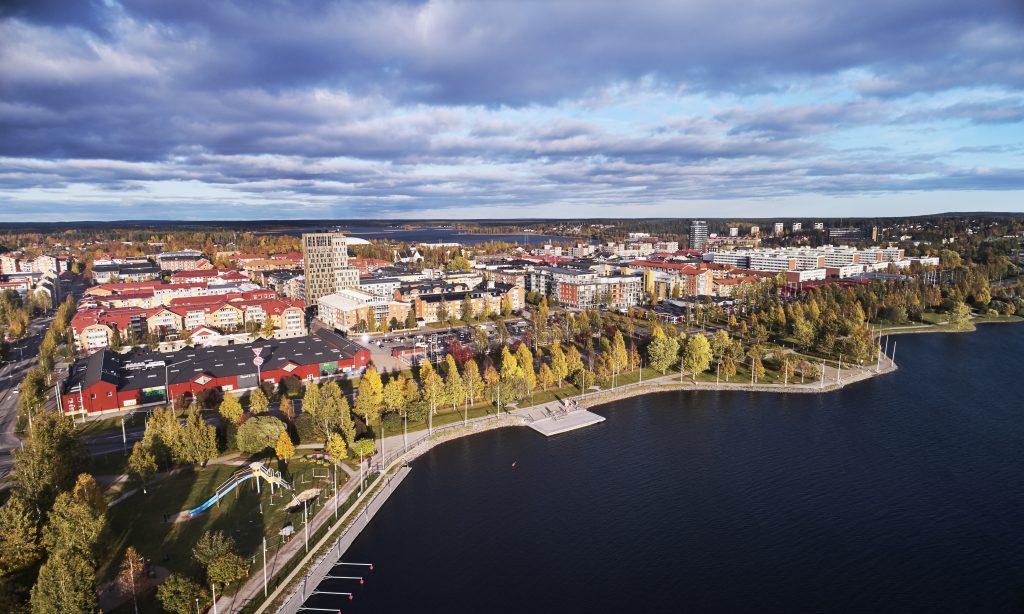 Ett drönarfoto taget över en stad på hösten. Träden i stadens parker och längs gatorna har färger som varierar från gyllene gult till varmt rött, vilket skapar ett färgrikt mönster bland byggnaderna. Himlen är klarblå men fylld av dramatiska, mjukt formade moln som ger en känsla av rörelse och djup. Solens ljus faller över stadens tak och gator, och lyfter fram byggnadernas konturer och höstens färger på ett mjukt, varmt sätt. Bilden ger en översikt över stadens struktur och naturens skiftningar samtidigt som ljus och moln skapar en stämningsfull atmosfär.