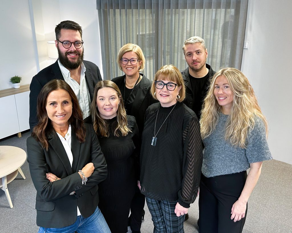 Gruppbild på sju personer inom inköp. Formellt men avslappnat klädda, står nära varandra och ler varmt. Positiv och välkomnande känsla.