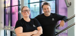 Två personer i svarta tröjor med samma logotyp står i en ljus trappa och ler. Färgat glas bakom dem skapar ett mjukt, modernt intryck.