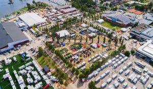 Flygbild över ett livligt mässområde i Piteå med tält, maskiner och husbilar. Människor rör sig mellan montrar, intill vatten, grönområden och stadskvarter.