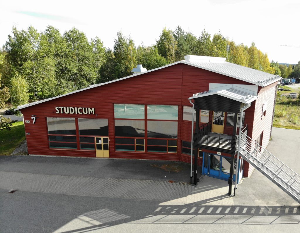 Röd byggnad med stora fönster och skylten Studicum på fasaden. Entré med trappa och ramp. Huset ligger nära Acusticum, omgivet av träd.