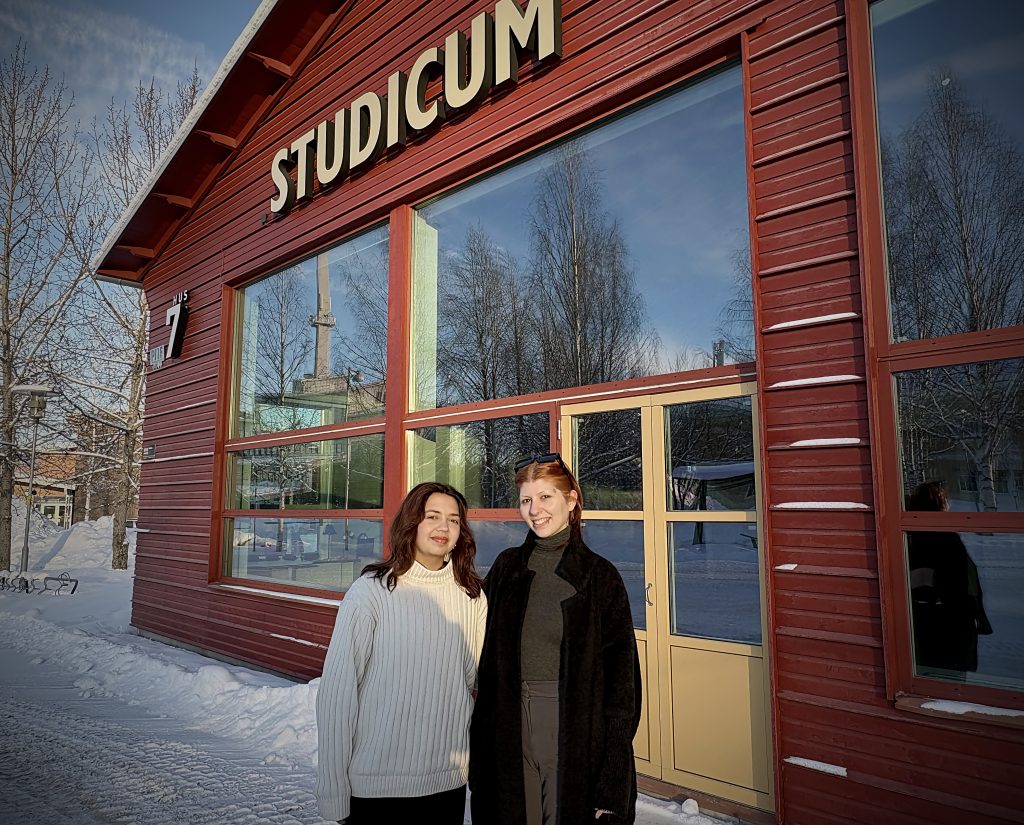 Två kvinnor står utanför en röd byggnad med skylten “Studicum”. Snö ligger på marken och kvällssolen lyser varmt mot fasaden.